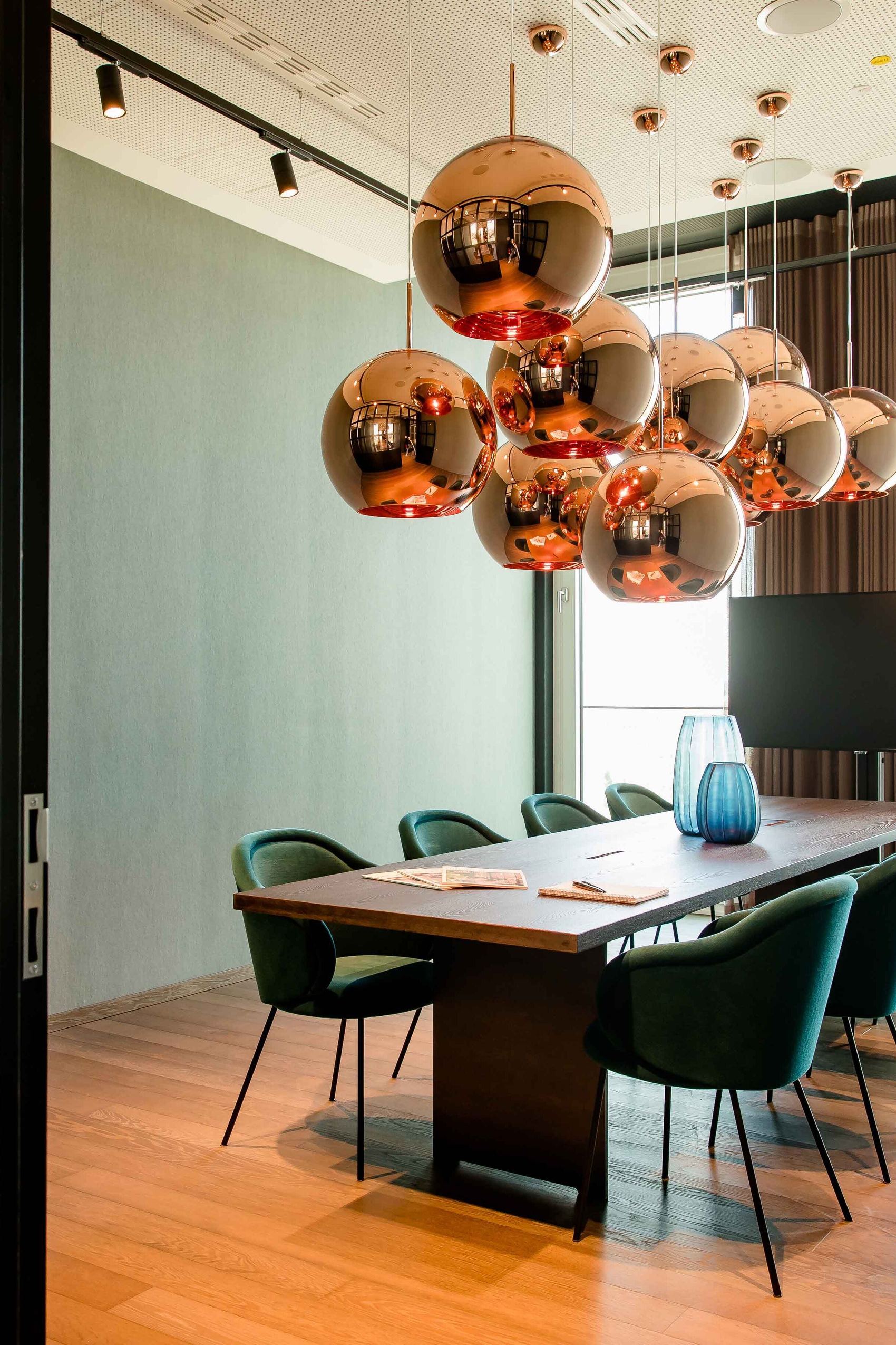 Motel One Konferenzraum mit langem Holztisch, acht grünen Stühlen und kupferfarbenen Kugelpendelleuchten, hell durch großflächige Fenster mit Tageslicht erleuchtet.