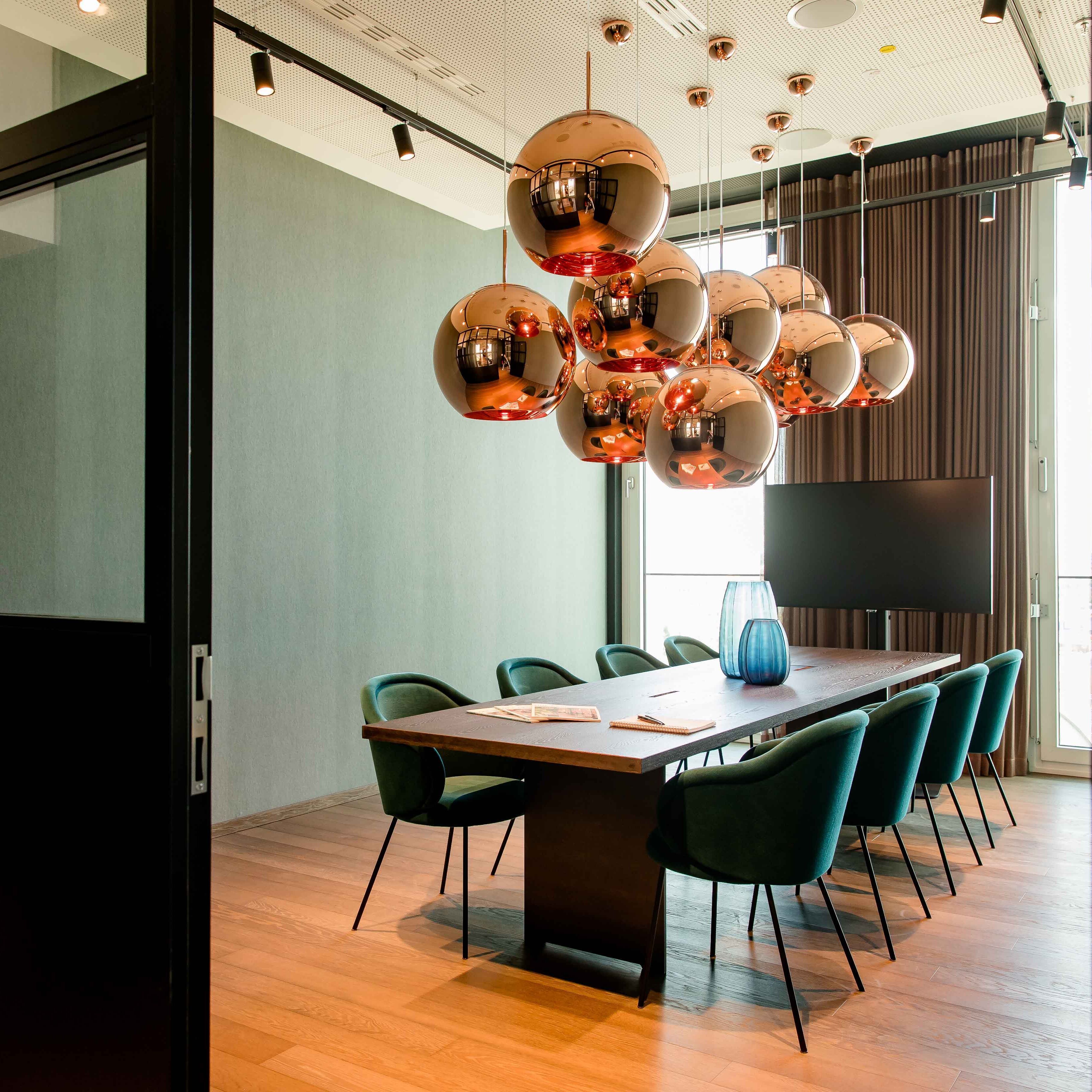 Motel One Konferenzraum mit langem Holztisch, acht grünen Stühlen und kupferfarbenen Kugelpendelleuchten, hell durch großflächige Fenster mit Tageslicht erleuchtet.