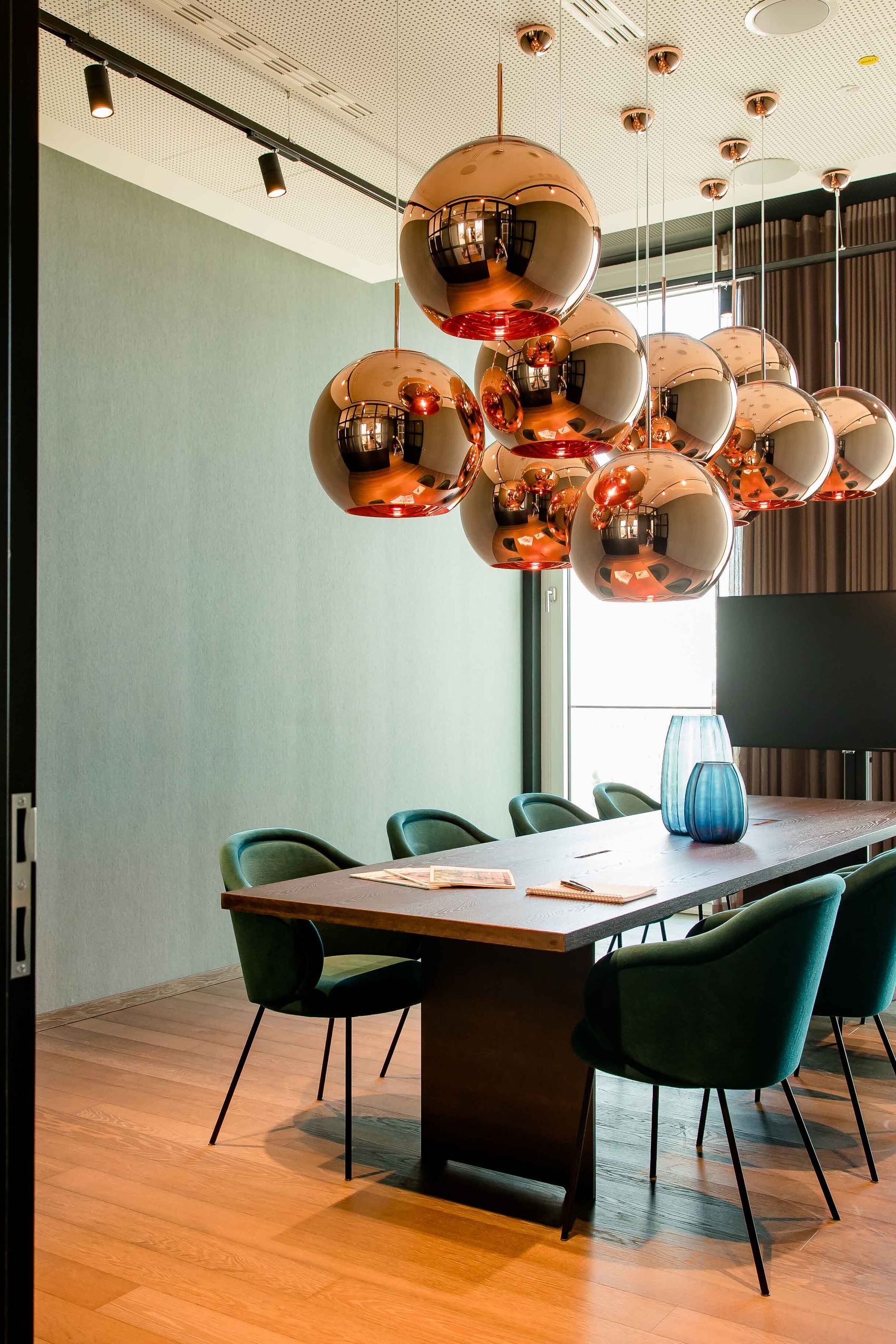 Motel One Konferenzraum mit langem Holztisch, acht grünen Stühlen und kupferfarbenen Kugelpendelleuchten, hell durch großflächige Fenster mit Tageslicht erleuchtet.