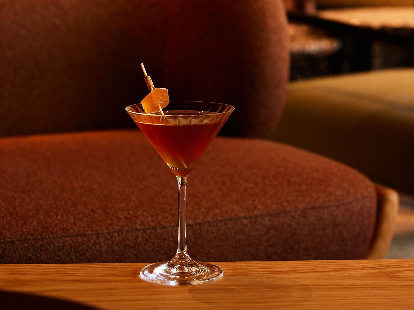 Ein eleganter Cocktail in einem Martiniglas steht auf einem Holztisch vor einer gemütlichen, braunen Sofa- und Sitzgruppe in einer stilvollen Hotelbar-Atmosphäre.