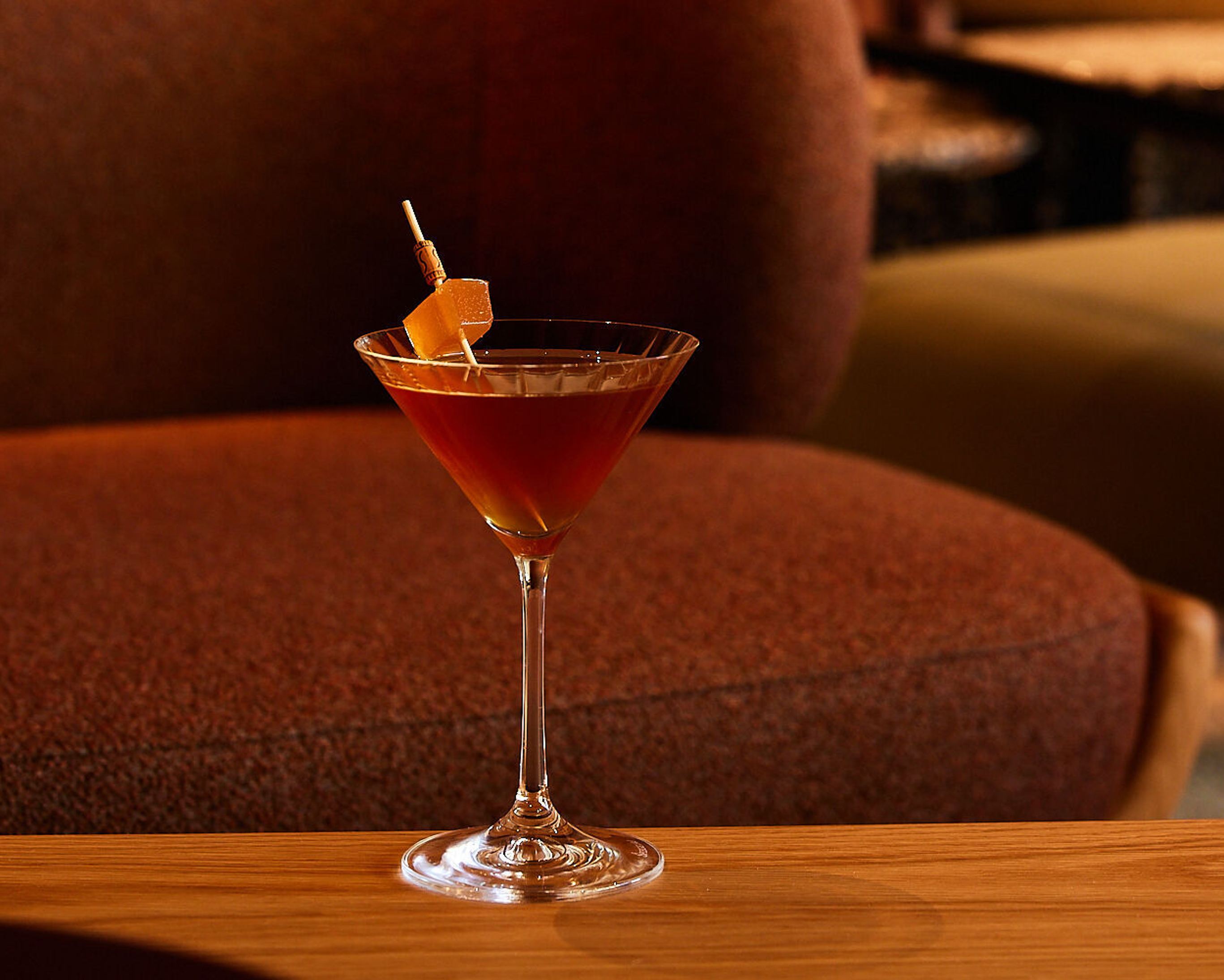 Ein eleganter Cocktail in einem Martiniglas steht auf einem Holztisch vor einer gemütlichen, braunen Sofa- und Sitzgruppe in einer stilvollen Hotelbar-Atmosphäre.