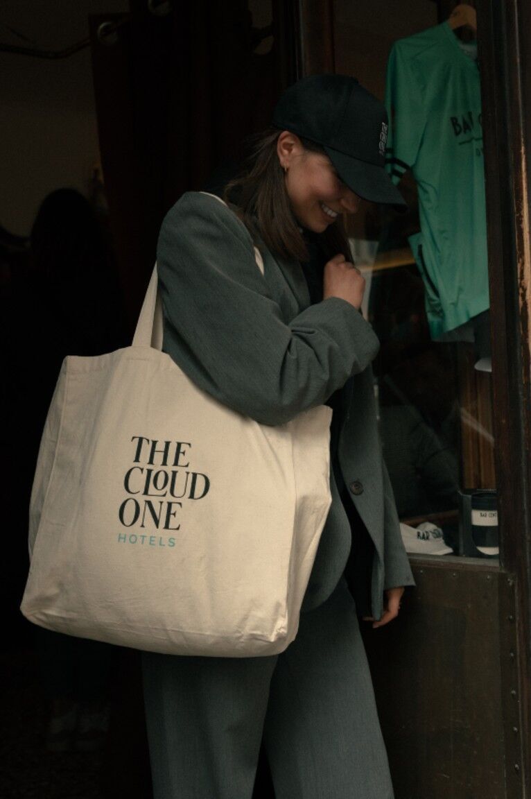 Eine lächelnde Frau in dunkelgrüner Kleidung trägt eine große Stofftasche mit dem Schriftzug „THE CLOUD ONE HOTELS“, die auf einen entspannten, modernen Hotelaufenthalt hinweist.