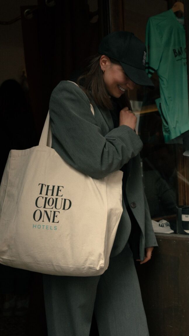 Eine lächelnde Frau in dunkelgrüner Kleidung trägt eine große Stofftasche mit dem Schriftzug „THE CLOUD ONE HOTELS“, die auf einen entspannten, modernen Hotelaufenthalt hinweist.