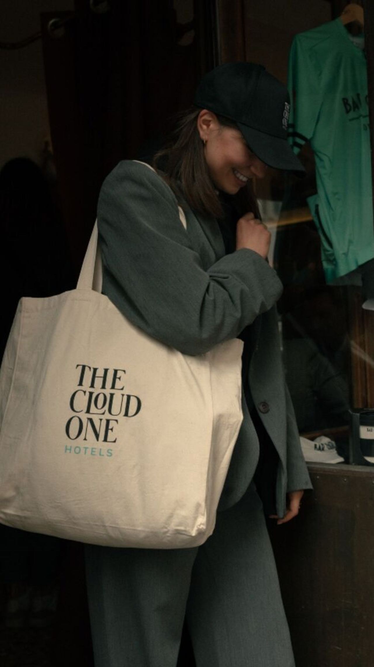 Eine lächelnde Frau in dunkelgrüner Kleidung trägt eine große Stofftasche mit dem Schriftzug „THE CLOUD ONE HOTELS“, die auf einen entspannten, modernen Hotelaufenthalt hinweist. Eine lächelnde Frau in dunkelgrüner Kleidung trägt eine große Stofftasche mit dem Schriftzug „THE CLOUD ONE HOTELS“, die auf einen entspannten, modernen Hotelaufenthalt hinweist.