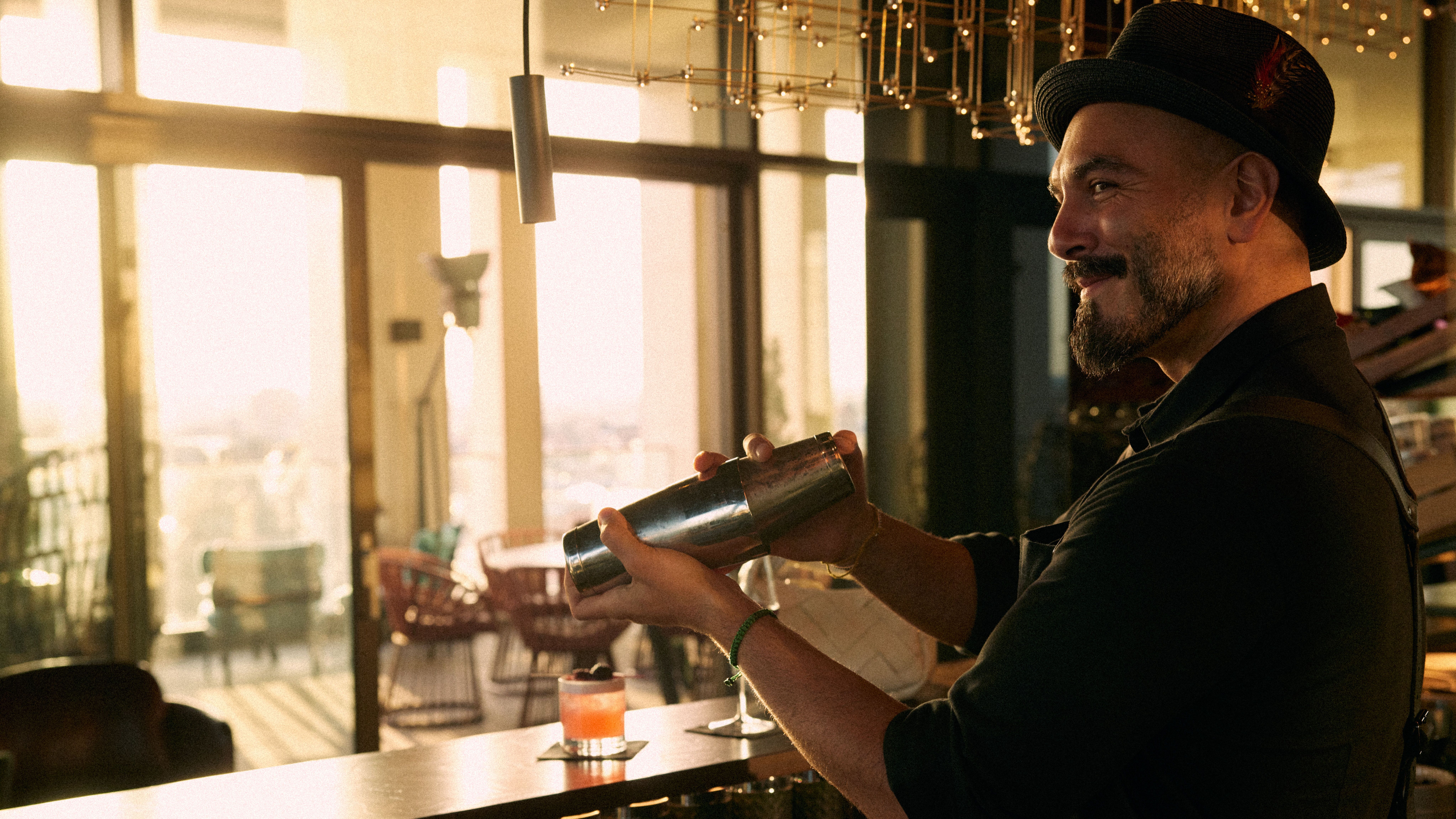 Barkeeper mixt mit einem Shaker einen Cocktail in stilvoller Bar mit großem Fenster und warmem, einladendem Licht am Abend.