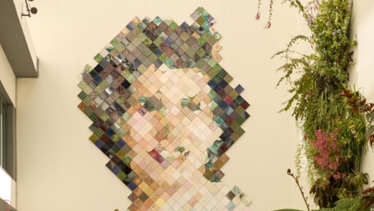Großes Wandkunstwerk aus bunten Quadraten, das ein Frauenportrait darstellt, im hellen, modernen Innenhof mit Sitzgelegenheiten und üppigen grünen Pflanzen.