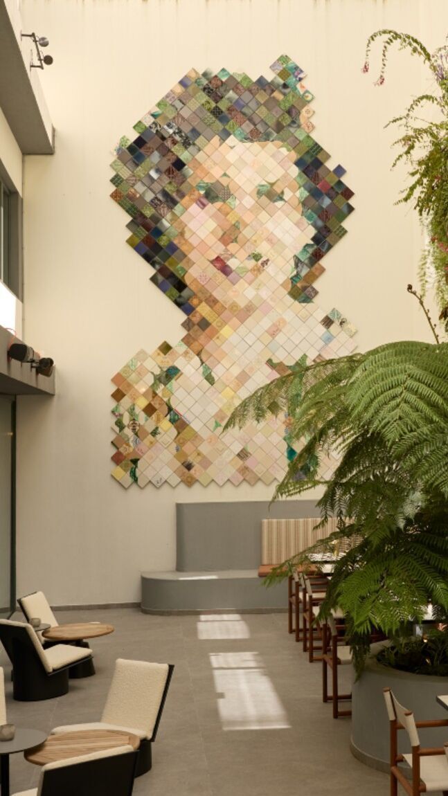 Großes Wandkunstwerk aus bunten Quadraten, das ein Frauenportrait darstellt, im hellen, modernen Innenhof mit Sitzgelegenheiten und üppigen grünen Pflanzen.