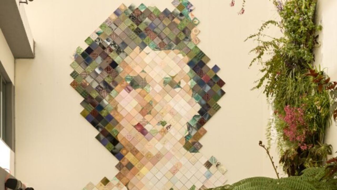 Großes Wandkunstwerk aus bunten Quadraten, das ein Frauenportrait darstellt, im hellen, modernen Innenhof mit Sitzgelegenheiten und üppigen grünen Pflanzen.