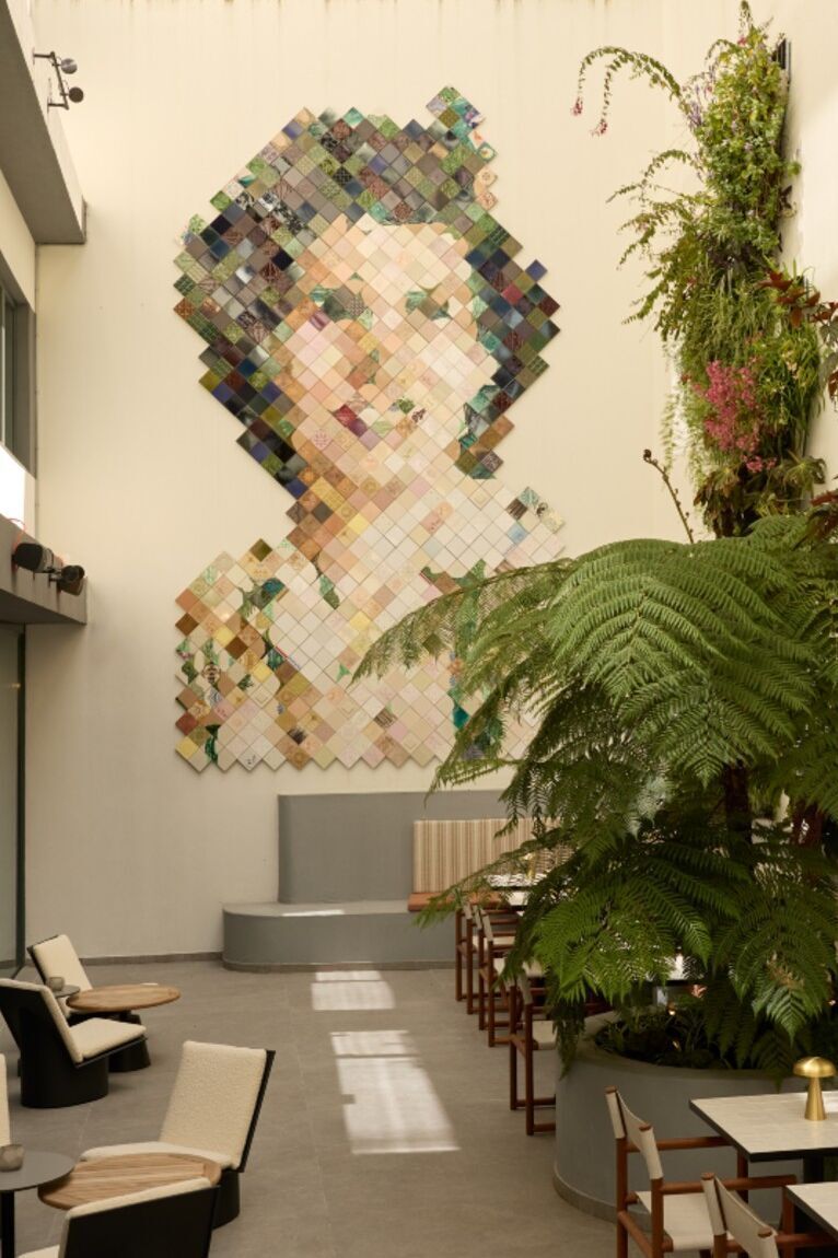 Großes Wandkunstwerk aus bunten Quadraten, das ein Frauenportrait darstellt, im hellen, modernen Innenhof mit Sitzgelegenheiten und üppigen grünen Pflanzen.