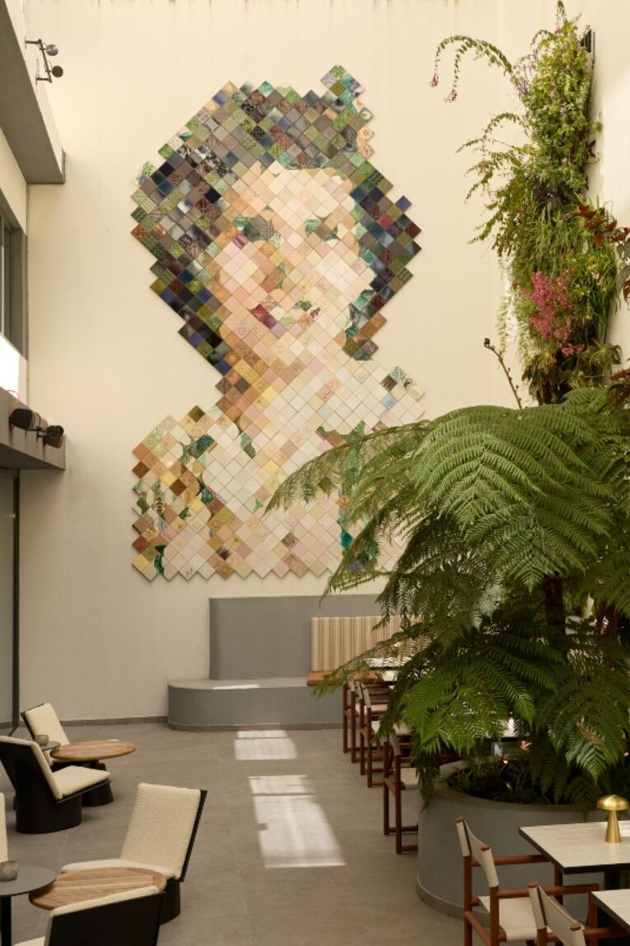 Großes Wandkunstwerk aus bunten Quadraten, das ein Frauenportrait darstellt, im hellen, modernen Innenhof mit Sitzgelegenheiten und üppigen grünen Pflanzen.