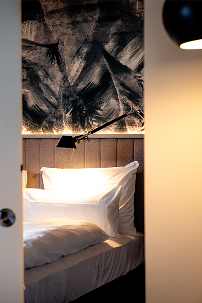 Blick durch geöffnete Zimmertür auf ein einladendes Bett mit weißen Kissen und Decke, sanfte Beleuchtung und moderne, künstlerische Wandgestaltung im Hintergrund. Blick durch geöffnete Zimmertür auf ein einladendes Bett mit weißen Kissen und Decke, sanfte Beleuchtung und moderne, künstlerische Wandgestaltung im Hintergrund.