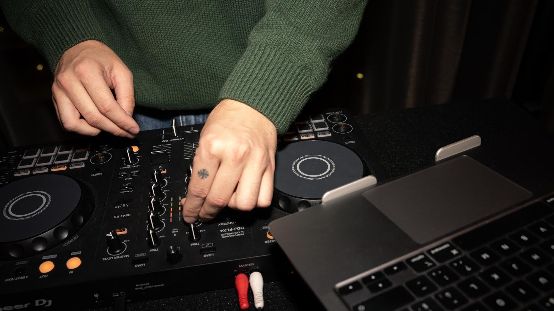 Hände eines DJs, die an einem Mischpult Musik einstellen, daneben ein geöffnetes Laptop, Atmosphäre kreativ und konzentriert.