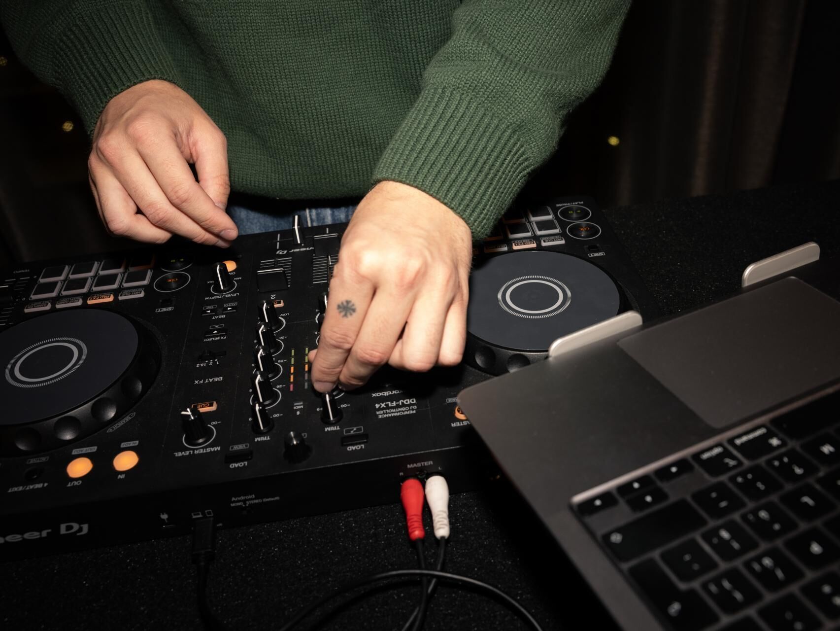 Hände eines DJs, die an einem Mischpult Musik einstellen, daneben ein geöffnetes Laptop, Atmosphäre kreativ und konzentriert.