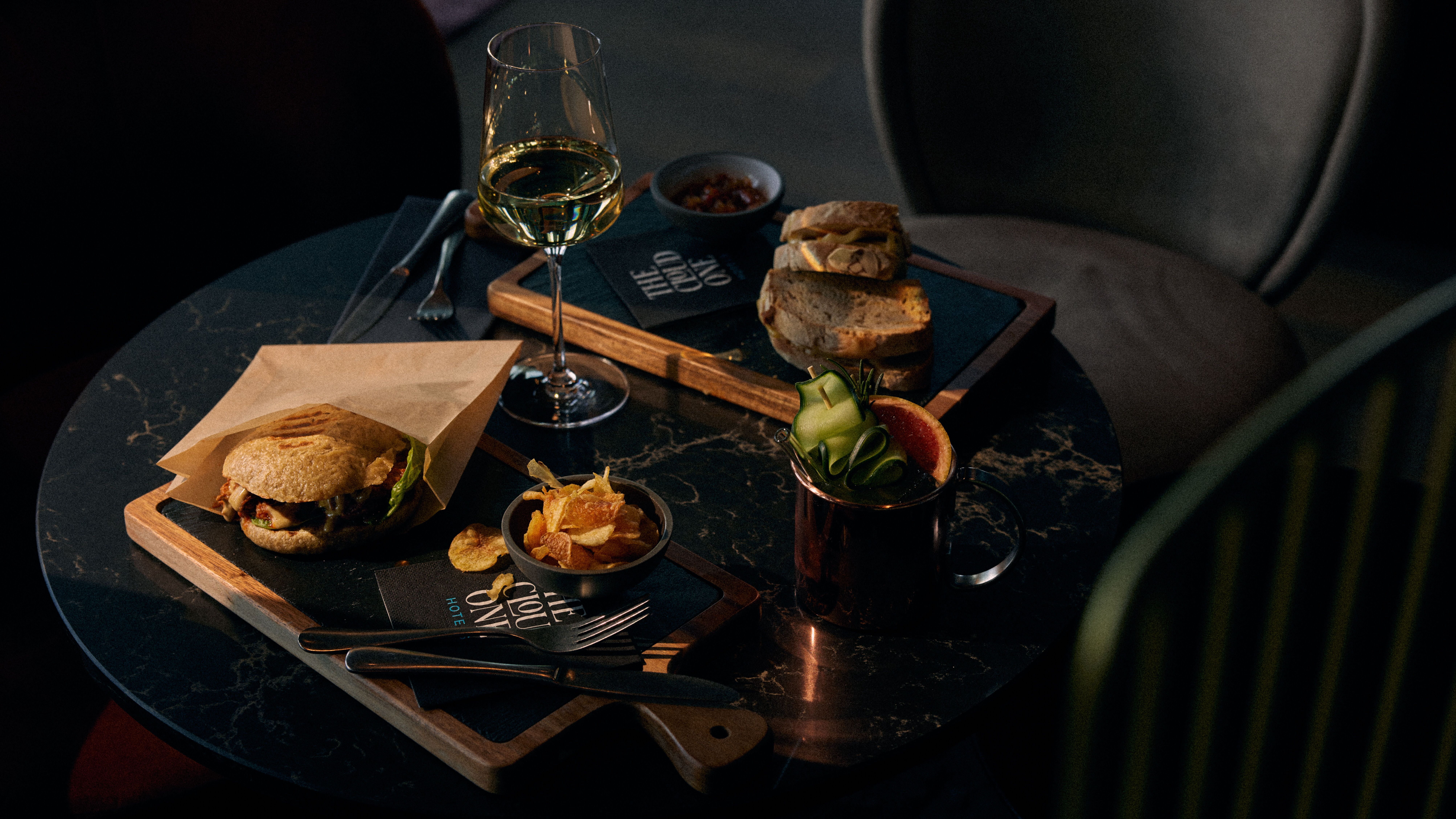 Auf einem dunklen Marmortisch stehen ein Burger mit Pommes, ein Glas Weißwein, frisches Brot und ein Cocktail mit Gurkenscheiben, arrangiert für einen genussvollen, entspannten Abend.