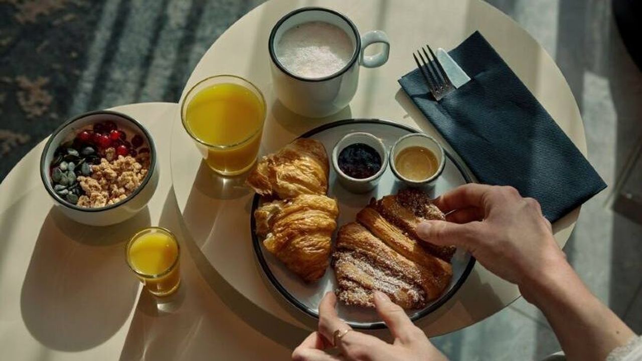 Frühstückstisch mit Croissants, Marmelade, Honig, zwei Gläsern Orangensaft, einer Tasse Kaffee und Schale mit Joghurt, Beeren und Müsli, Hände greifen nach Croissant.