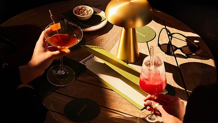 Vier Personen genießen bunte Getränke in stilvollen Gläsern an einem runden Holztisch, der von warmem Licht einer modernen Lampe beleuchtet wird. Die Atmosphäre wirkt entspannt und gesellig.