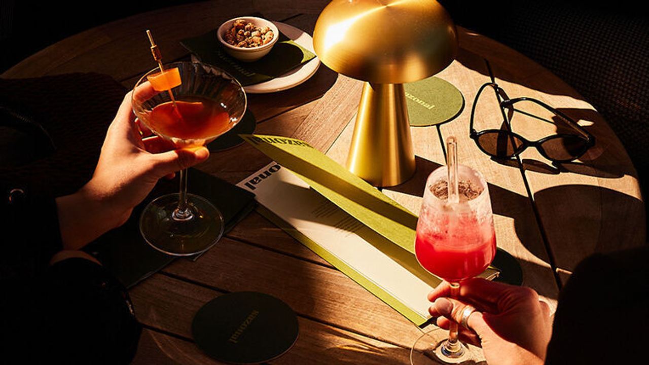 Vier Personen genießen bunte Getränke in stilvollen Gläsern an einem runden Holztisch, der von warmem Licht einer modernen Lampe beleuchtet wird. Die Atmosphäre wirkt entspannt und gesellig.