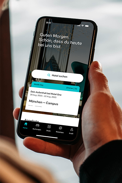 Hand hält Smartphone mit Hotel-App, zeigt Buchungsdetails für Motel One in München-Campus, inklusive Aufenthalt und Zimmernummer. Begrüßungstext „Guten Morgen, schön, dass du heute bei uns bist.“ sichtbar. Suchfunktion für Hotels ebenfalls dargestellt.