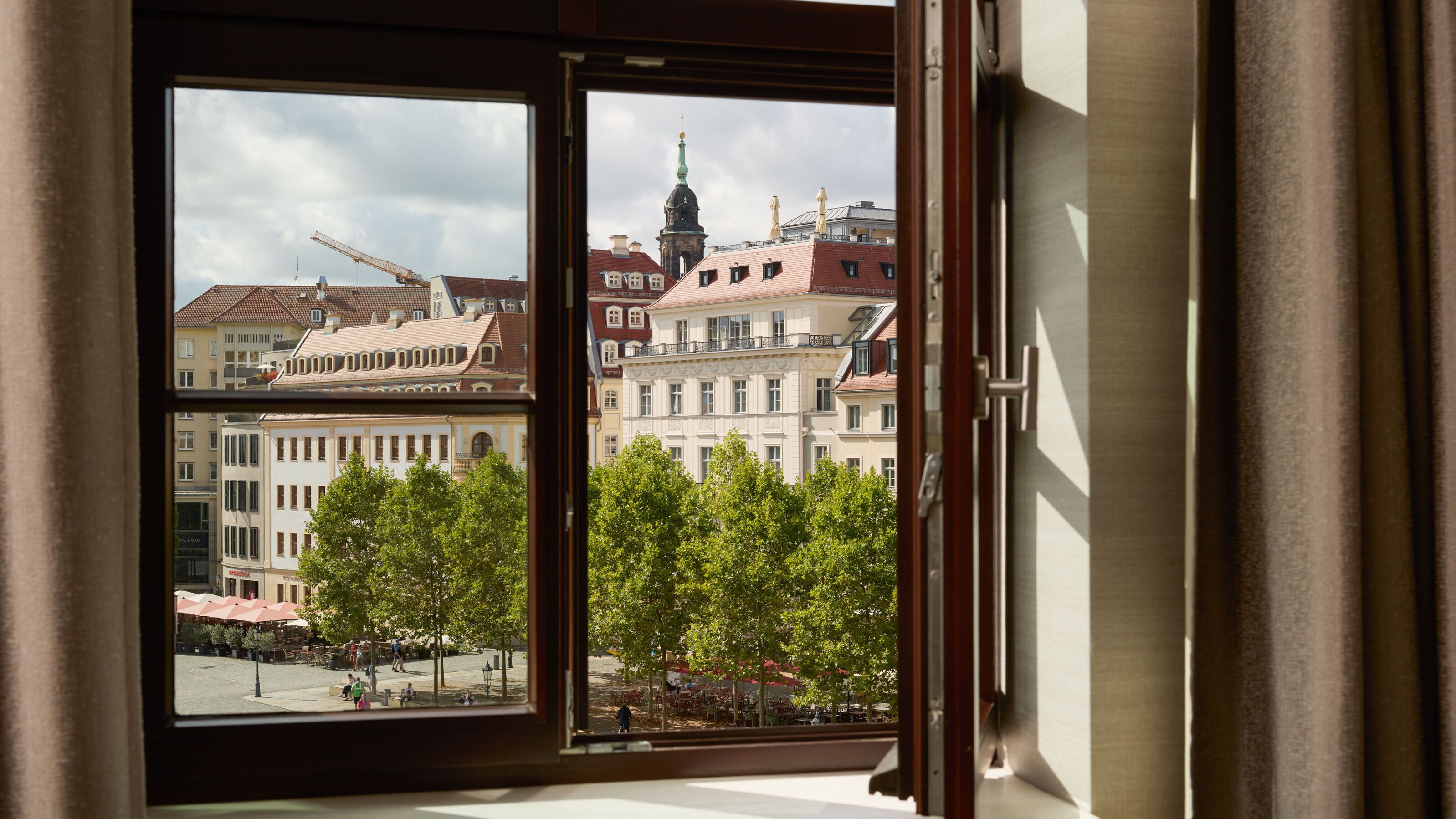 Blick aus offenem Hotelzimmerfenster auf historische Gebäude und grüne Bäume an einem städtischen Platz bei Tageslicht.