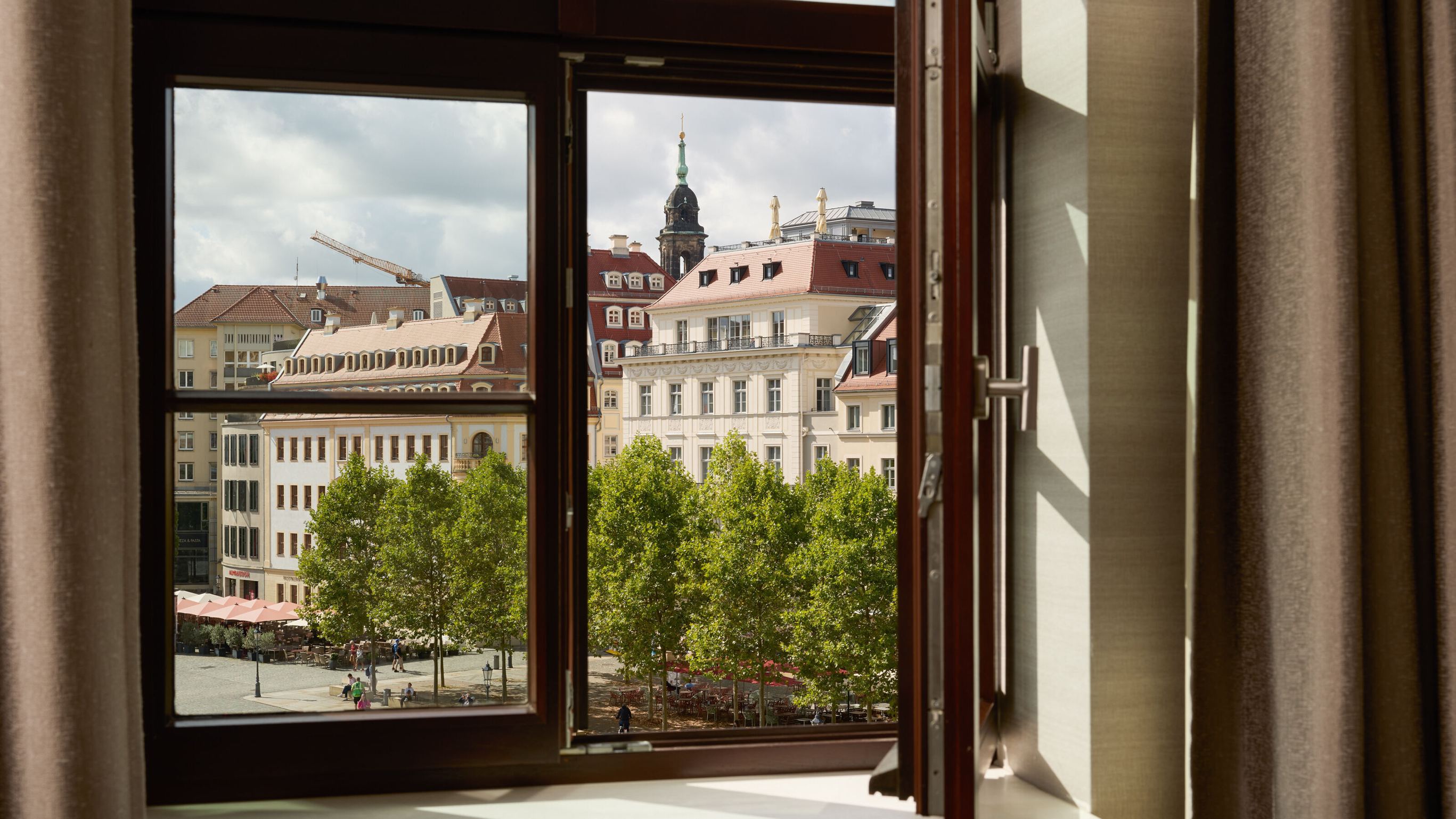 Blick aus offenem Hotelzimmerfenster auf historische Gebäude und grüne Bäume an einem städtischen Platz bei Tageslicht. Blick aus offenem Hotelzimmerfenster auf historische Gebäude und grüne Bäume an einem städtischen Platz bei Tageslicht.