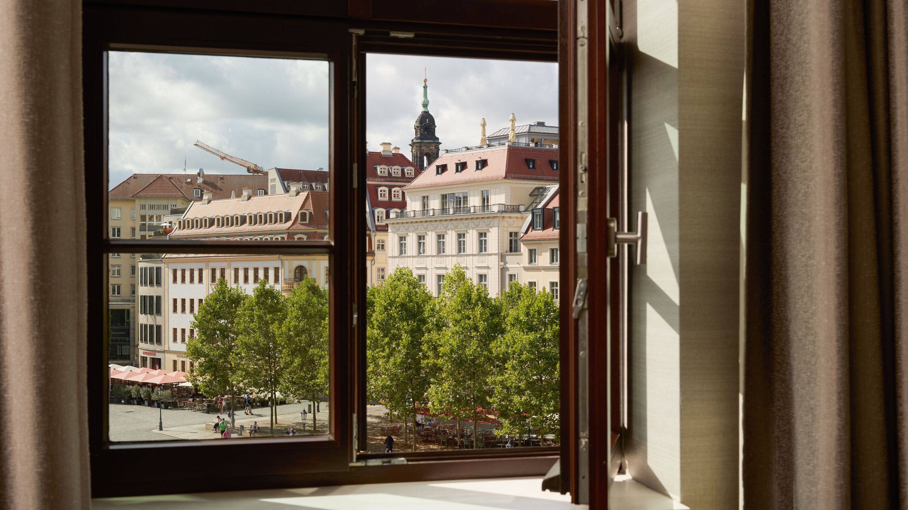 Blick aus offenem Hotelzimmerfenster auf historische Gebäude und grüne Bäume an einem städtischen Platz bei Tageslicht.
