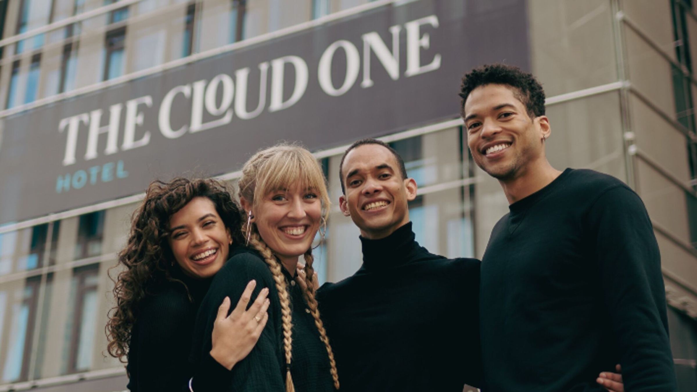 Vier junge Erwachsene stehen lachend vor dem Hotel "The Cloud One" und wirken entspannt und willkommen.