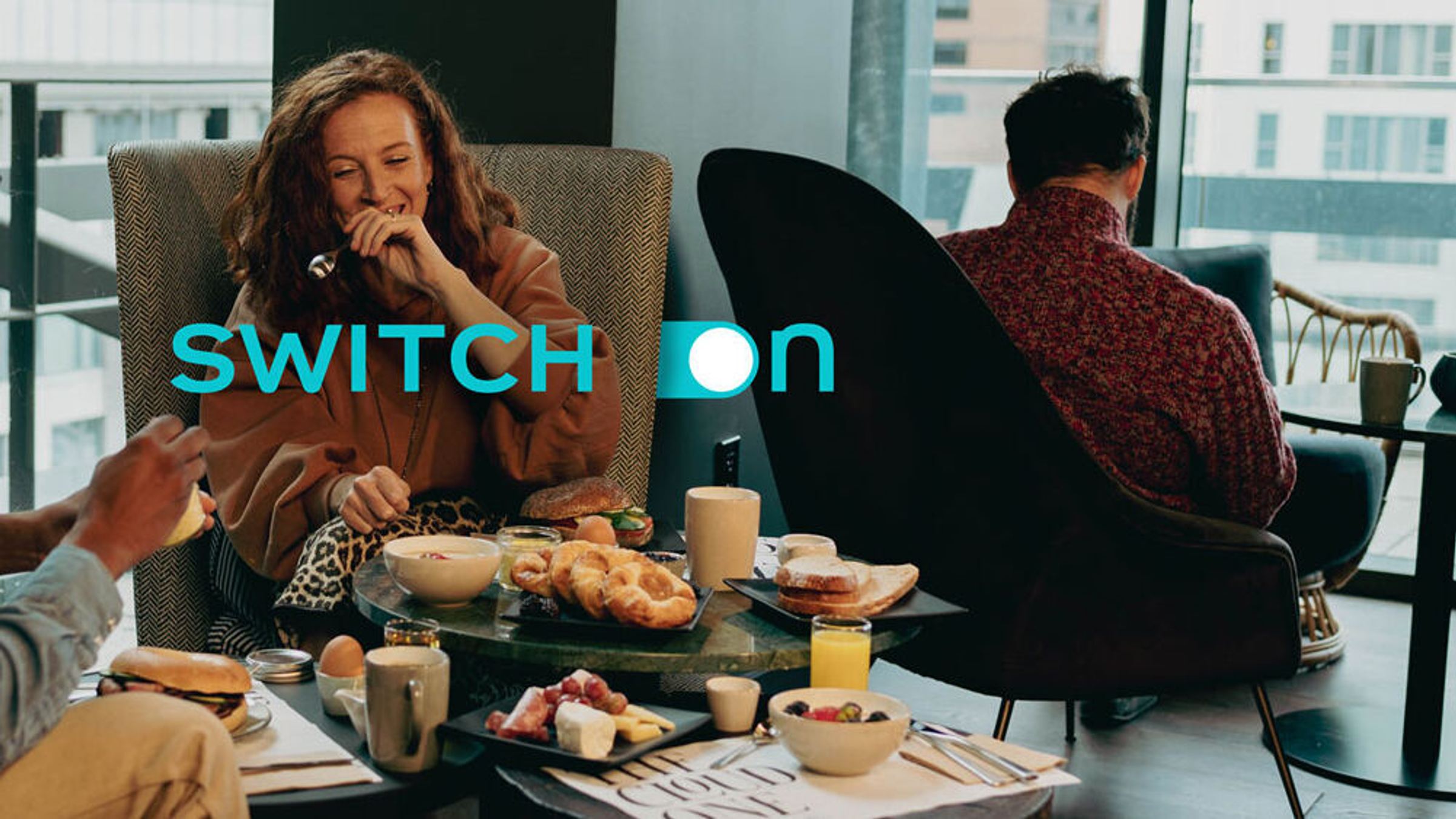 Gemütliche Frühstücksszene in Lounge-Bereich mit entspannten Gästen, Croissants, Getränken und Zeitungen, umgeben von großen Fenstern mit Stadtblick.