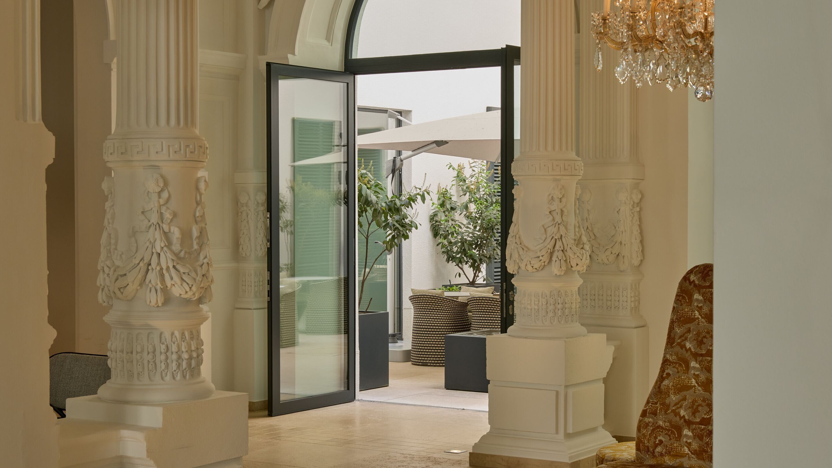 Heller, eleganter Innenraum mit prächtigen, verzierten Säulen und offenem Eingang zu einem Außenbereich mit Pflanzen und Sitzgelegenheiten unter Sonnenschirmen.
