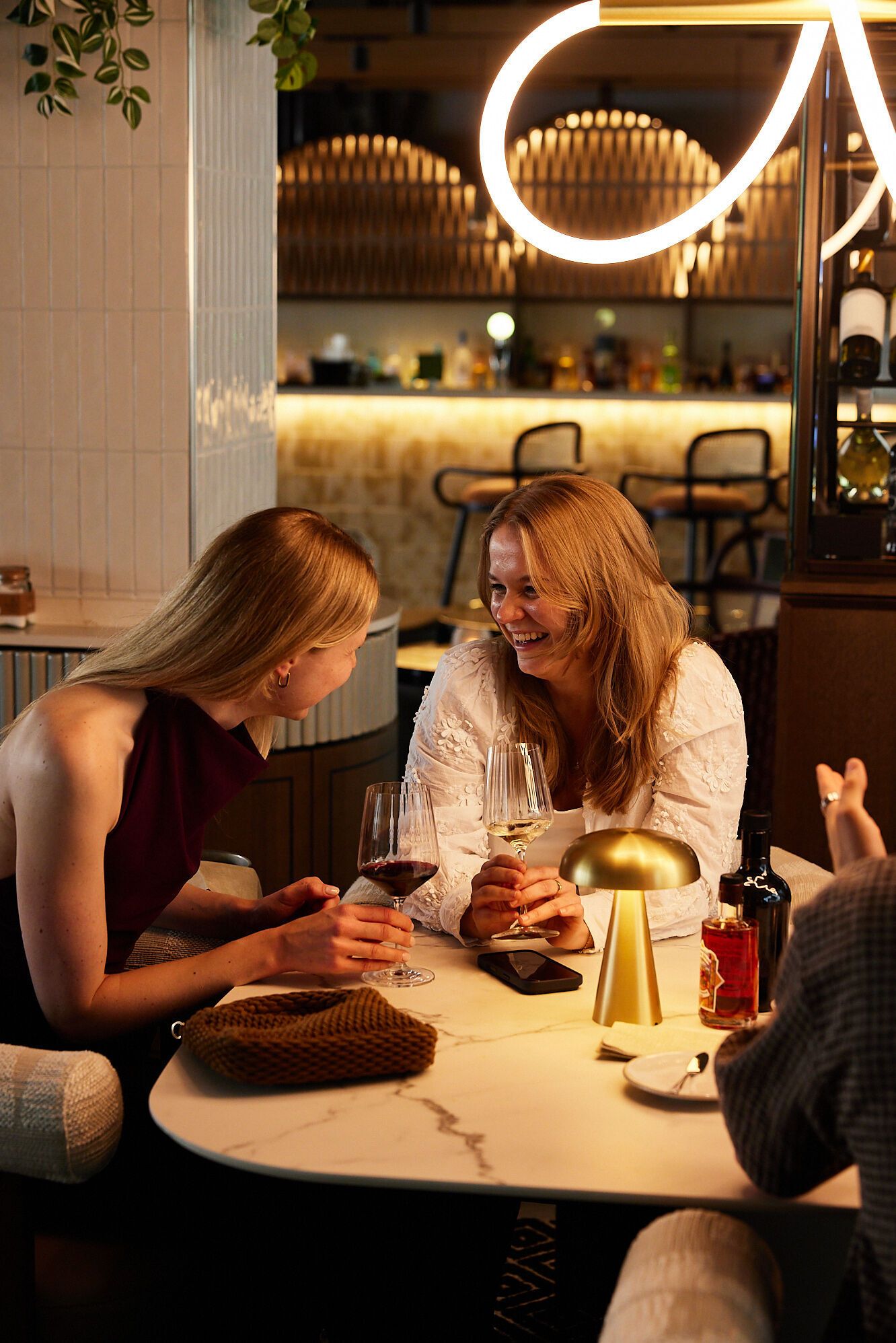 Zwei Frauen sitzen in gemütlicher Baratmosphäre, lachen und genießen Wein bei gedämpftem Licht und elegantem Ambiente, was gesellige und entspannte Momente vermittelt.