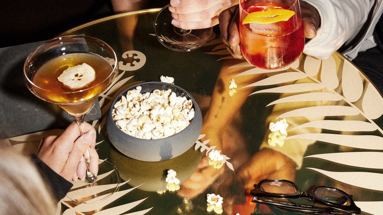 Drei Personen sitzen an einem Tisch, halten unterschiedliche Getränke – ein Glas Weißwein, einen Cocktail und ein Aperitif – und teilen eine Schüssel mit Popcorn. Die Atmosphäre wirkt entspannt und gesellig. Drei Personen sitzen an einem Tisch, halten unterschiedliche Getränke – ein Glas Weißwein, einen Cocktail und ein Aperitif – und teilen eine Schüssel mit Popcorn. Die Atmosphäre wirkt entspannt und gesellig.