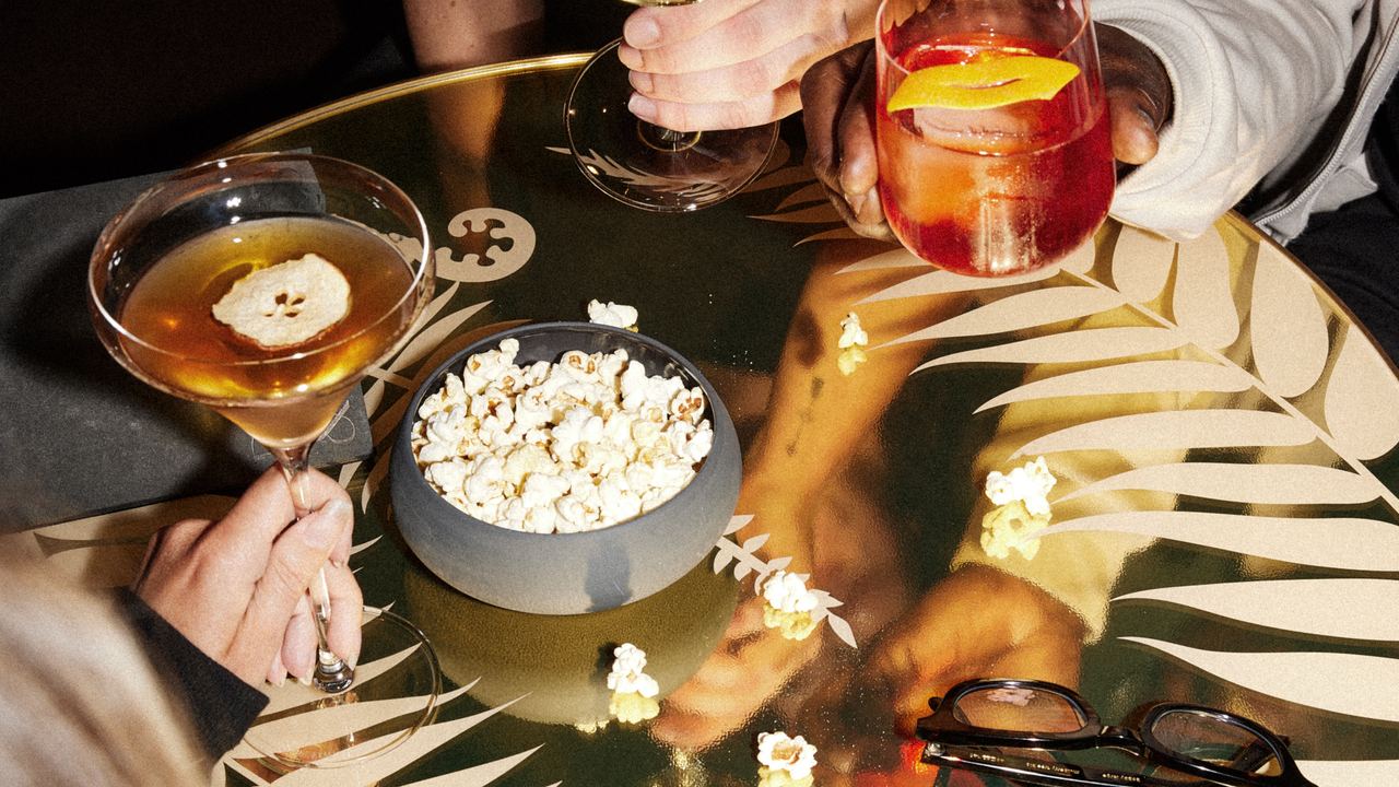 Drei Personen sitzen an einem Tisch, halten unterschiedliche Getränke – ein Glas Weißwein, einen Cocktail und ein Aperitif – und teilen eine Schüssel mit Popcorn. Die Atmosphäre wirkt entspannt und gesellig.