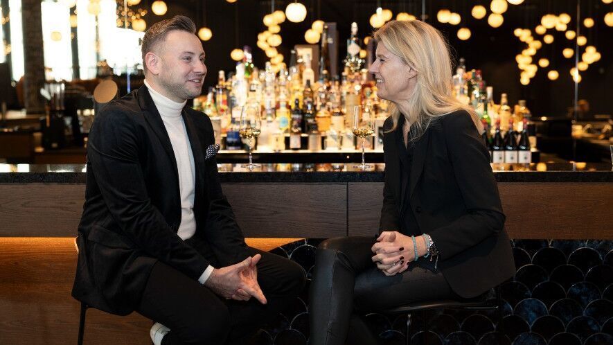 Zwei Gäste unterhalten sich entspannt an einer stilvollen Hotelbar mit warmem Licht und gut bestücktem Getränkeangebot im Hintergrund.