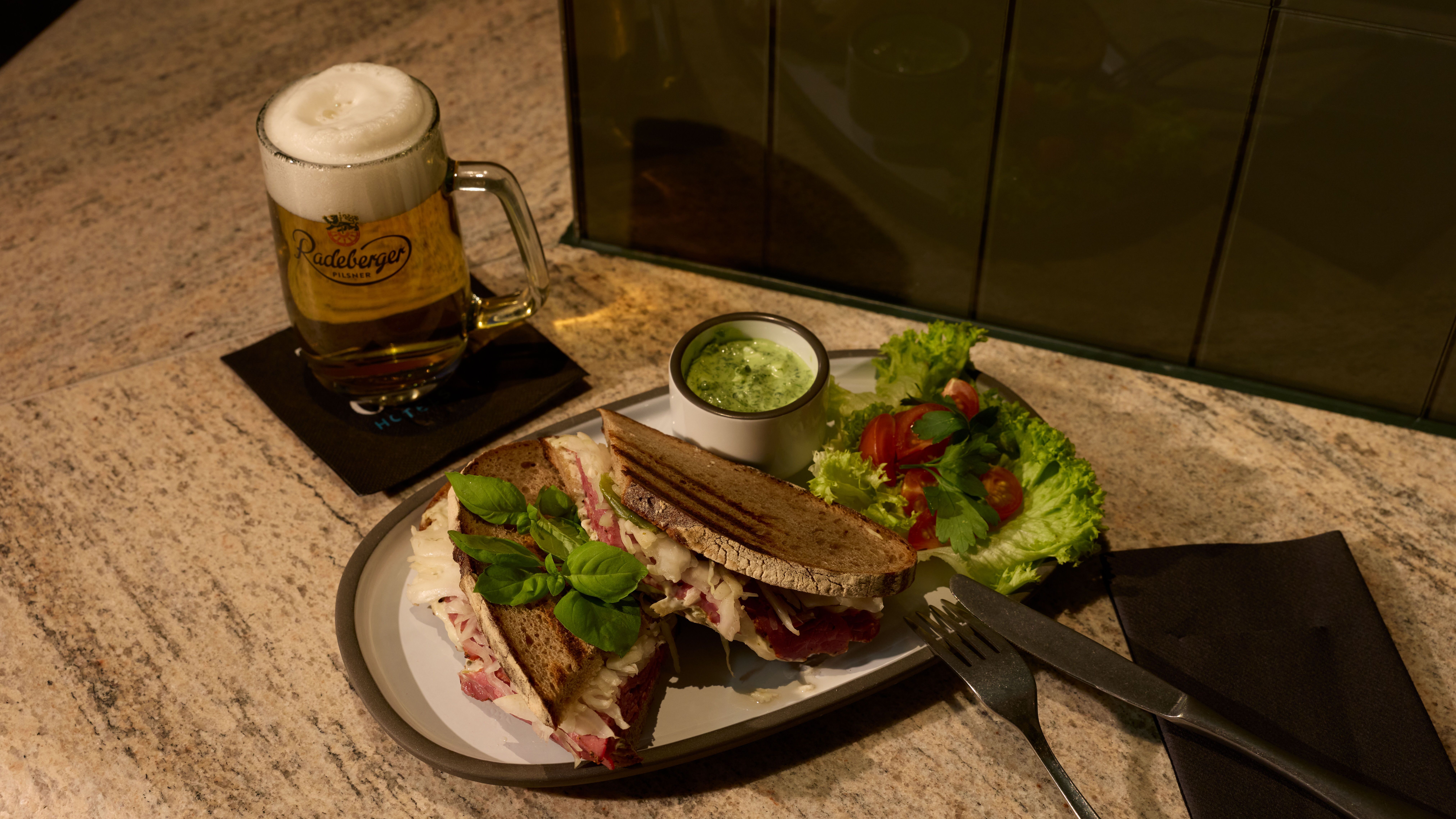 Sandwich mit frischem Salat und Dip auf Teller, daneben ein Glas Bier mit Schaum, serviert auf einem gedeckten Tisch.