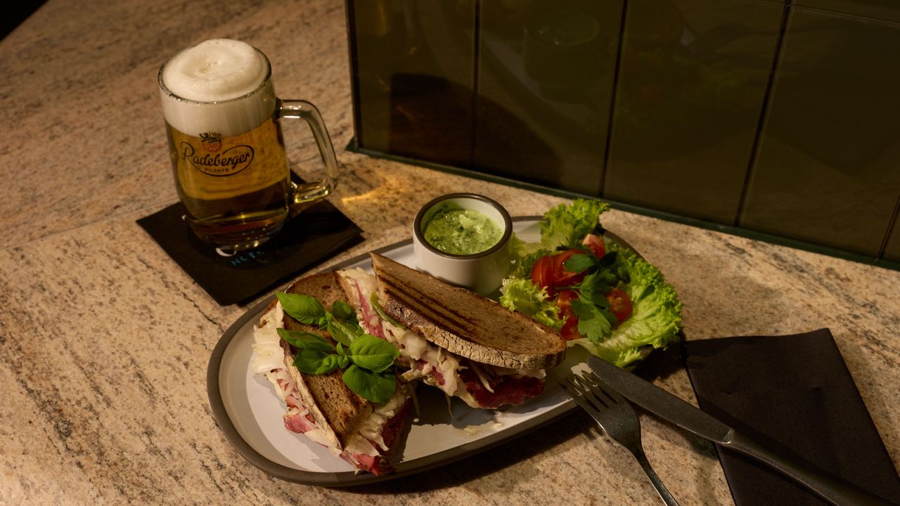 Sandwich mit frischem Salat und Dip auf Teller, daneben ein Glas Bier mit Schaum, serviert auf einem gedeckten Tisch. Sandwich mit frischem Salat und Dip auf Teller, daneben ein Glas Bier mit Schaum, serviert auf einem gedeckten Tisch.