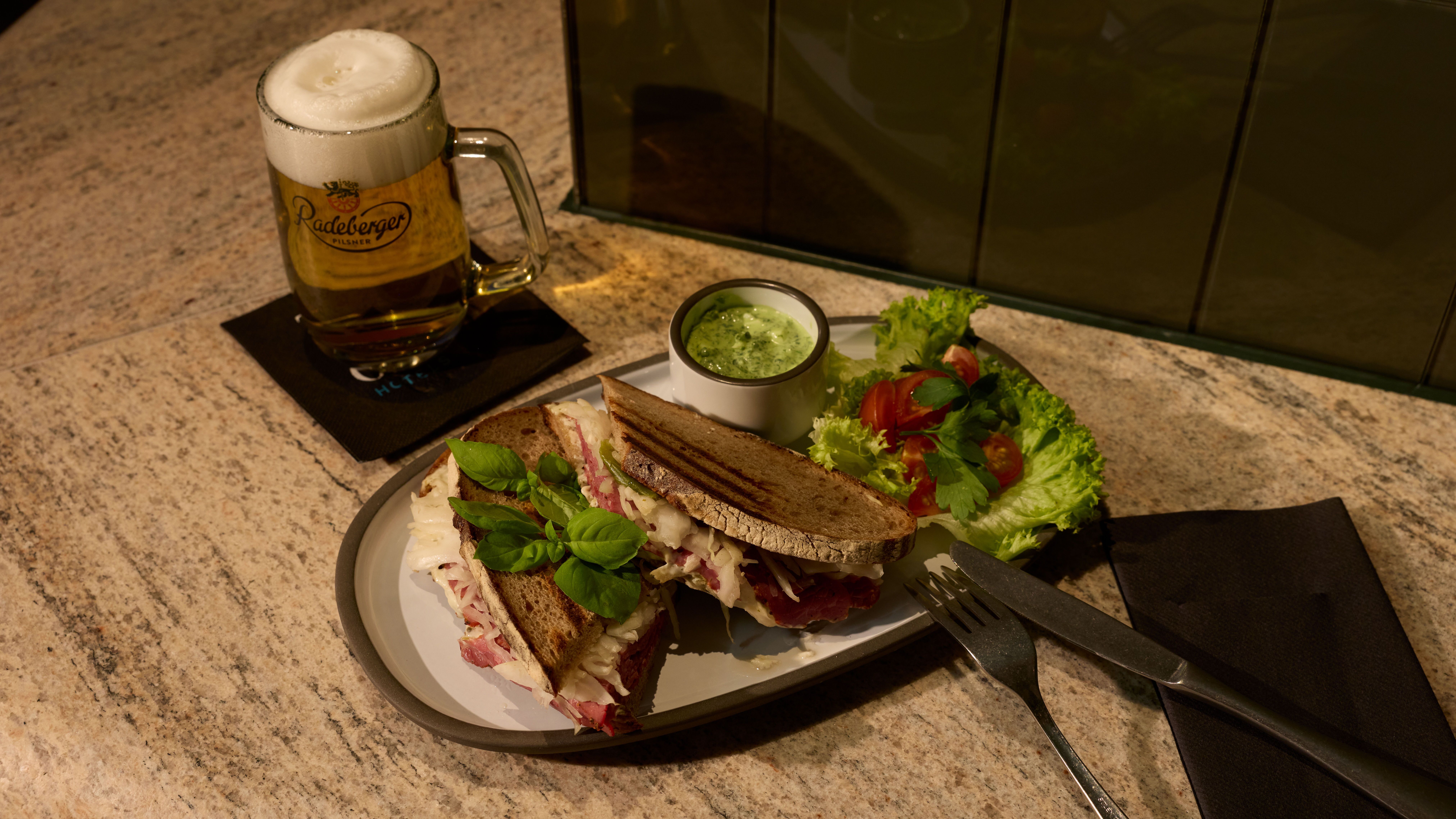 Sandwich mit frischem Salat und Dip auf Teller, daneben ein Glas Bier mit Schaum, serviert auf einem gedeckten Tisch.