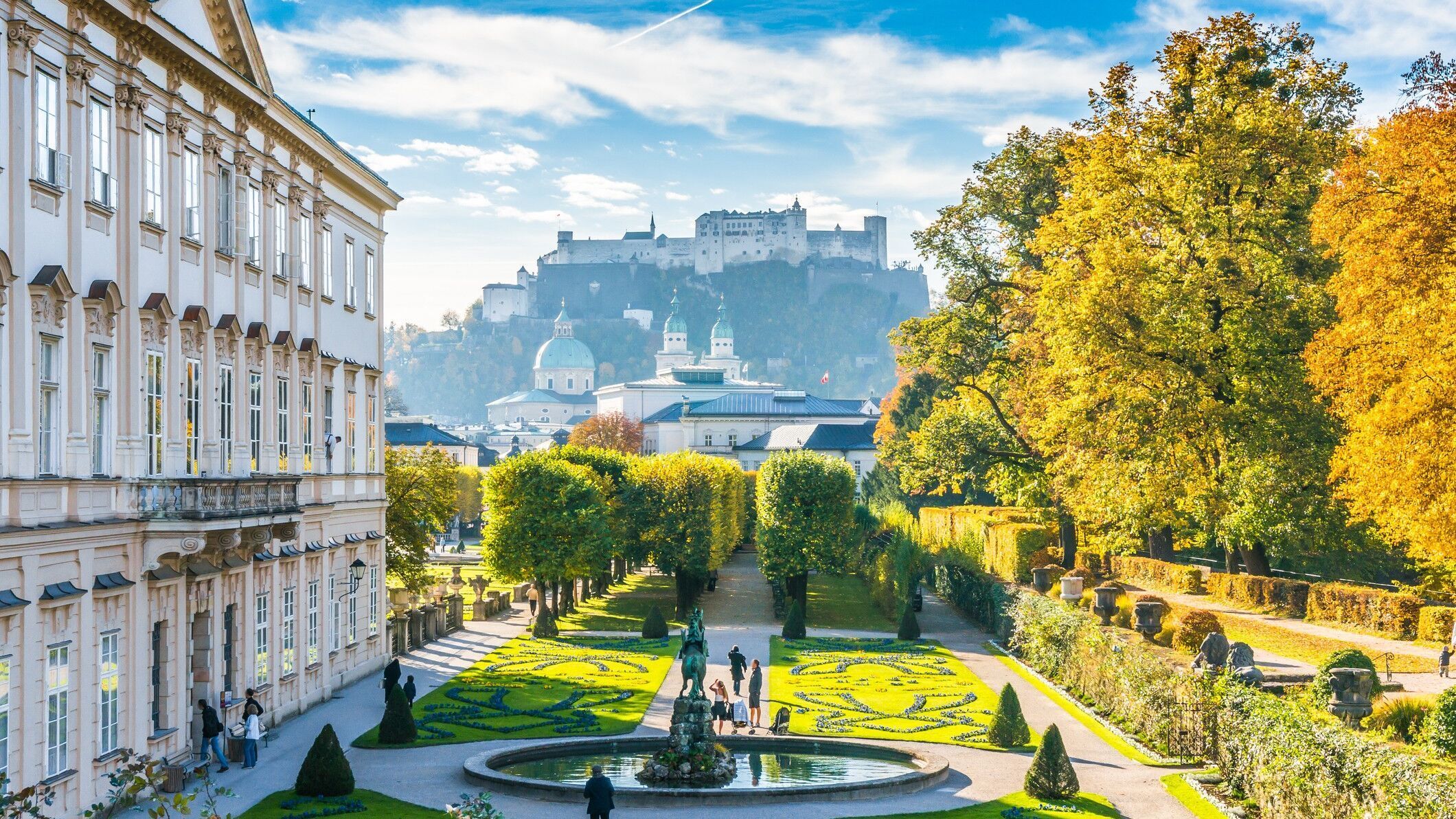Blick auf einen gepflegten Barockgarten mit kunstvoll geschnittenen Hecken und Springbrunnen, umgeben von historischen Gebäuden, mit der Festung Hohensalzburg im Hintergrund unter blauem Himmel.