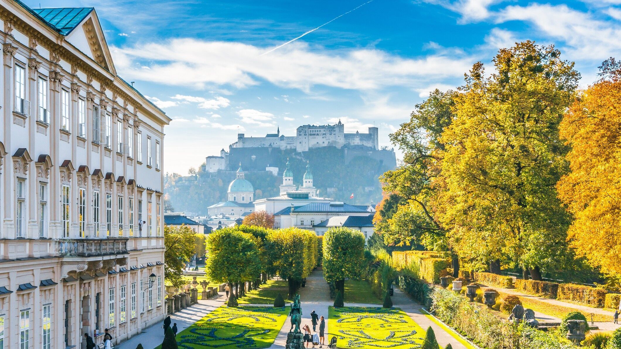 Blick auf einen gepflegten Barockgarten mit kunstvoll geschnittenen Hecken und Springbrunnen, umgeben von historischen Gebäuden, mit der Festung Hohensalzburg im Hintergrund unter blauem Himmel.