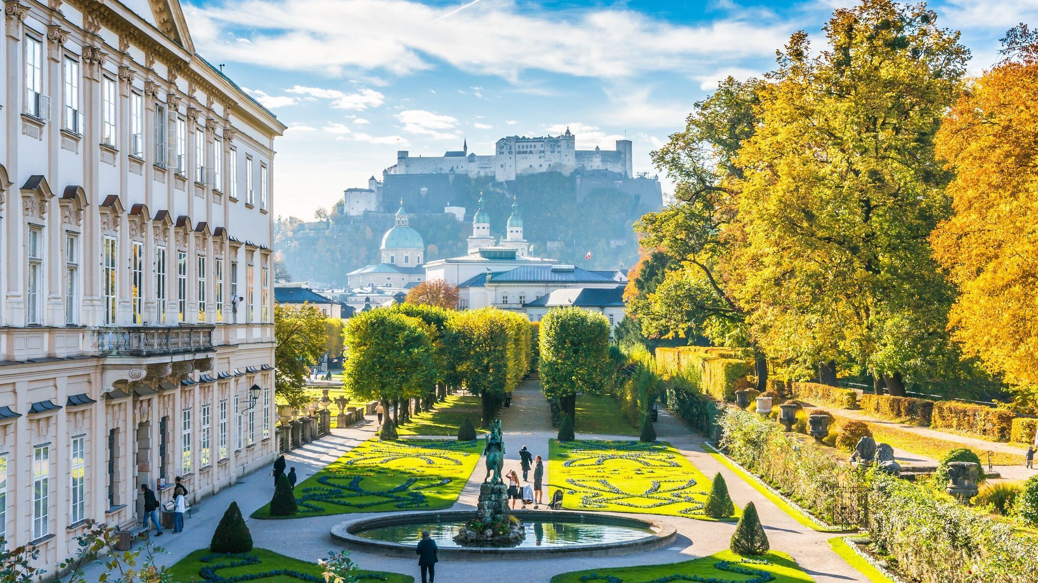 Blick auf einen gepflegten Barockgarten mit kunstvoll geschnittenen Hecken und Springbrunnen, umgeben von historischen Gebäuden, mit der Festung Hohensalzburg im Hintergrund unter blauem Himmel.