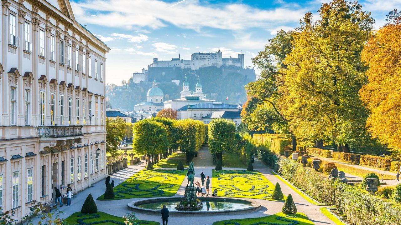 Blick auf einen gepflegten Barockgarten mit kunstvoll geschnittenen Hecken und Springbrunnen, umgeben von historischen Gebäuden, mit der Festung Hohensalzburg im Hintergrund unter blauem Himmel. Blick auf einen gepflegten Barockgarten mit kunstvoll geschnittenen Hecken und Springbrunnen, umgeben von historischen Gebäuden, mit der Festung Hohensalzburg im Hintergrund unter blauem Himmel.