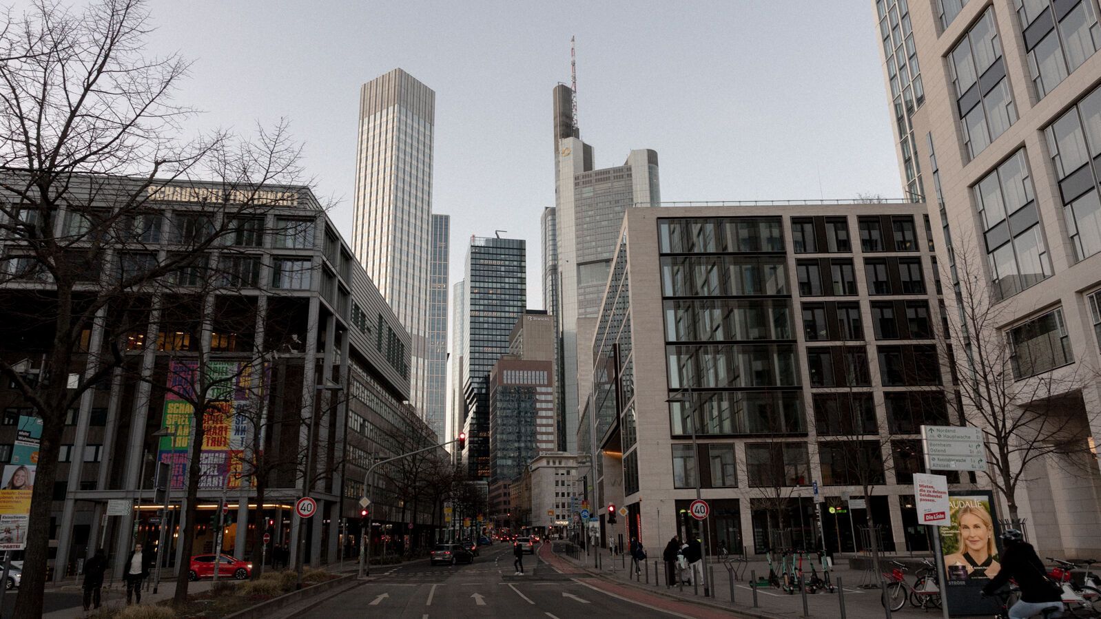 Straße in einem urbanen Geschäftsviertel mit modernen Hochhäusern und Bürogebäuden, vereinzelten Fußgängern und schwachem Tageslicht an einem bewölkten Tag.