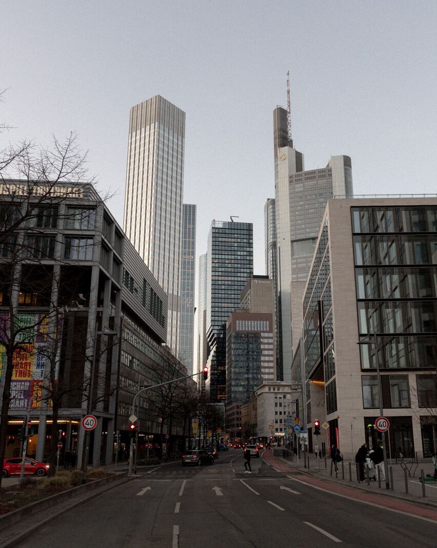 Straße in einem urbanen Geschäftsviertel mit modernen Hochhäusern und Bürogebäuden, vereinzelten Fußgängern und schwachem Tageslicht an einem bewölkten Tag.