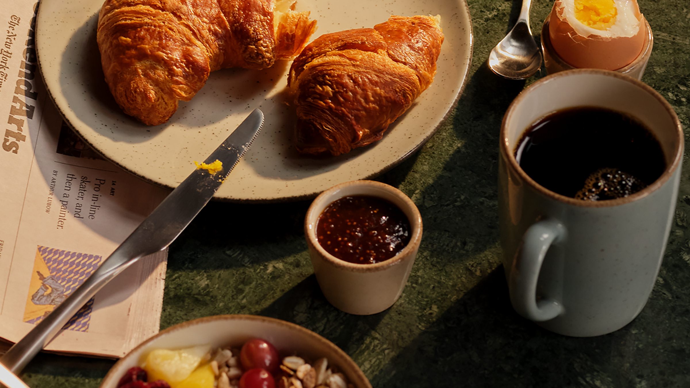 Frühstück mit frischen Croissants, weichem Ei, Kaffee und Marmelade auf grünem Tisch, begleitet von einer geöffneten Zeitung.