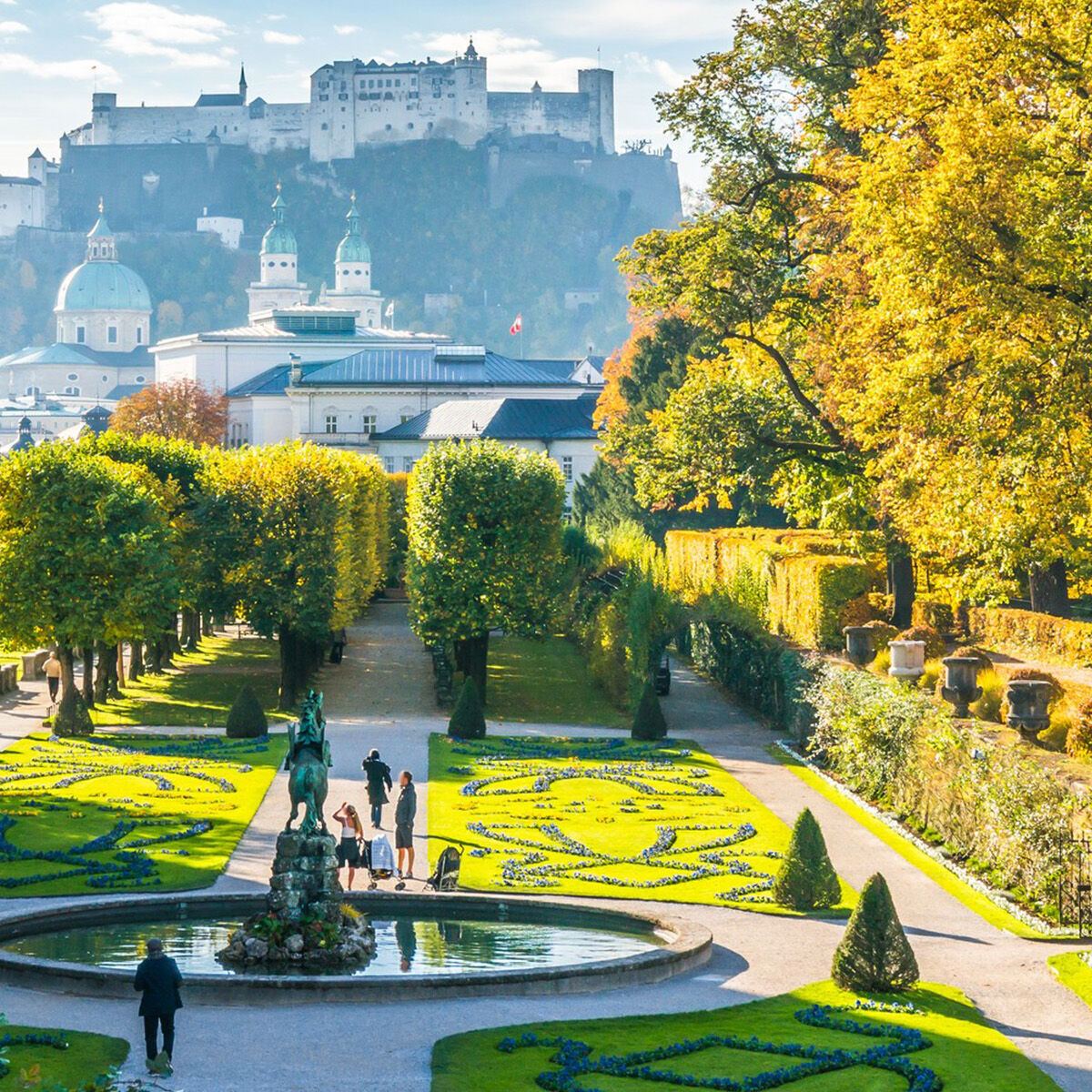 Blick auf den gepflegten Mirabellgarten mit Brunnen, Spaziergängern und herbstlich gefärbten Bäumen, im Hintergrund die Hohensalzburg Festung unter einem klaren Himmel.