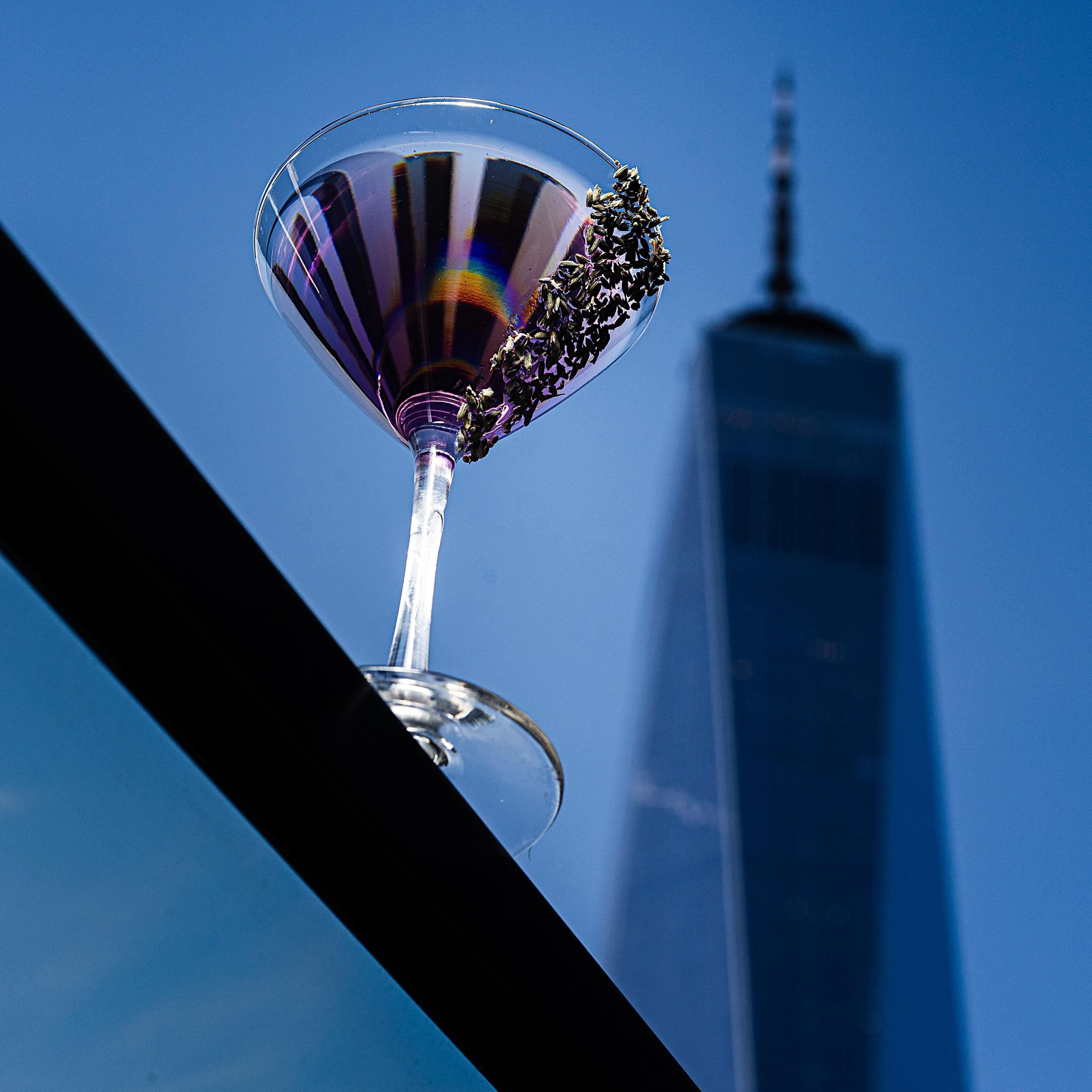Lilac Bliss Special Drink New York Sounds in the Cloud Cocktailglas mit einem lila Getränk und dekoriertem Glasrand vor blauem Himmel und Hochhaus-Silhouette.