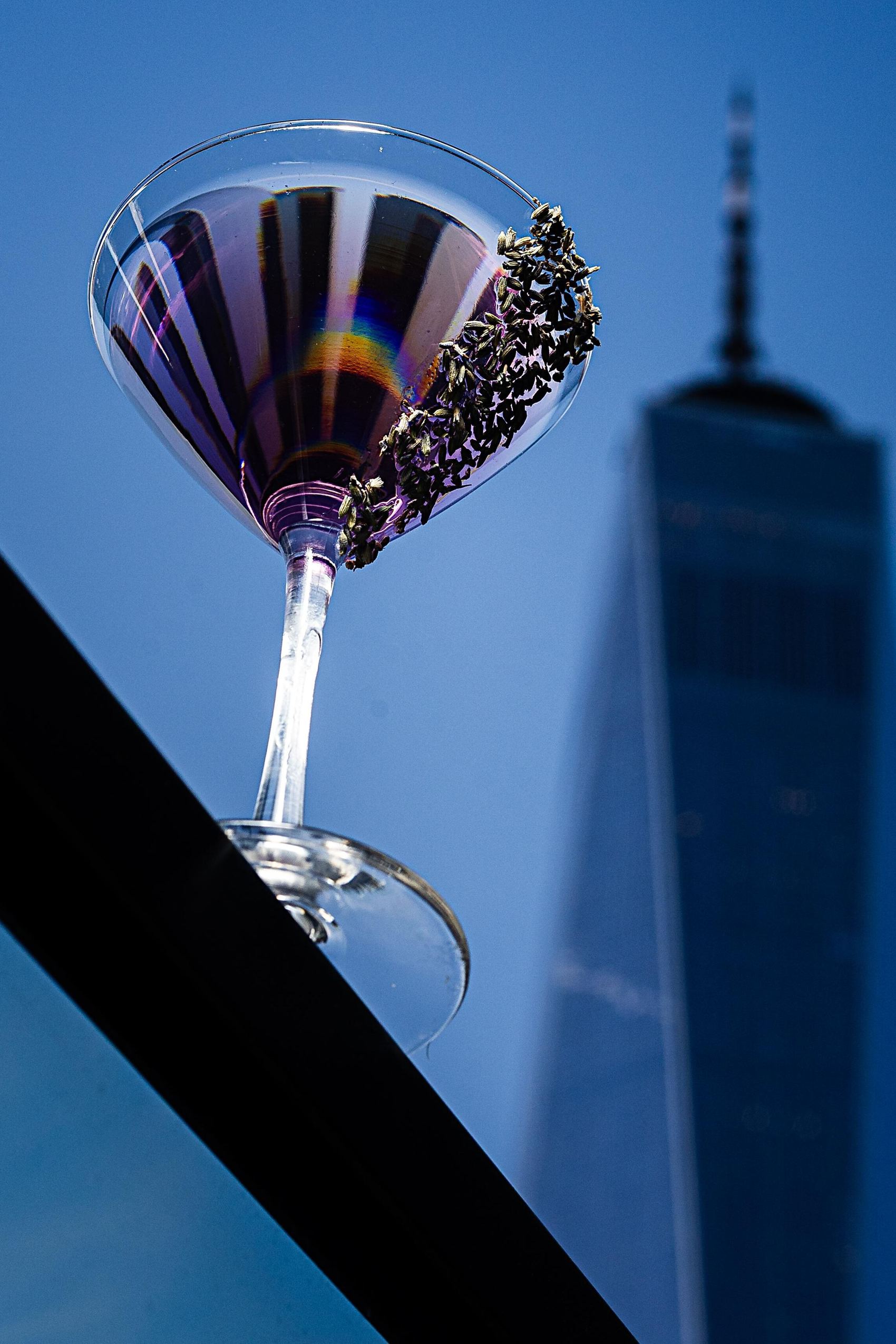 Lilac Bliss Special Drink New York Sounds in the Cloud Cocktailglas mit einem lila Getränk und dekoriertem Glasrand vor blauem Himmel und Hochhaus-Silhouette.
