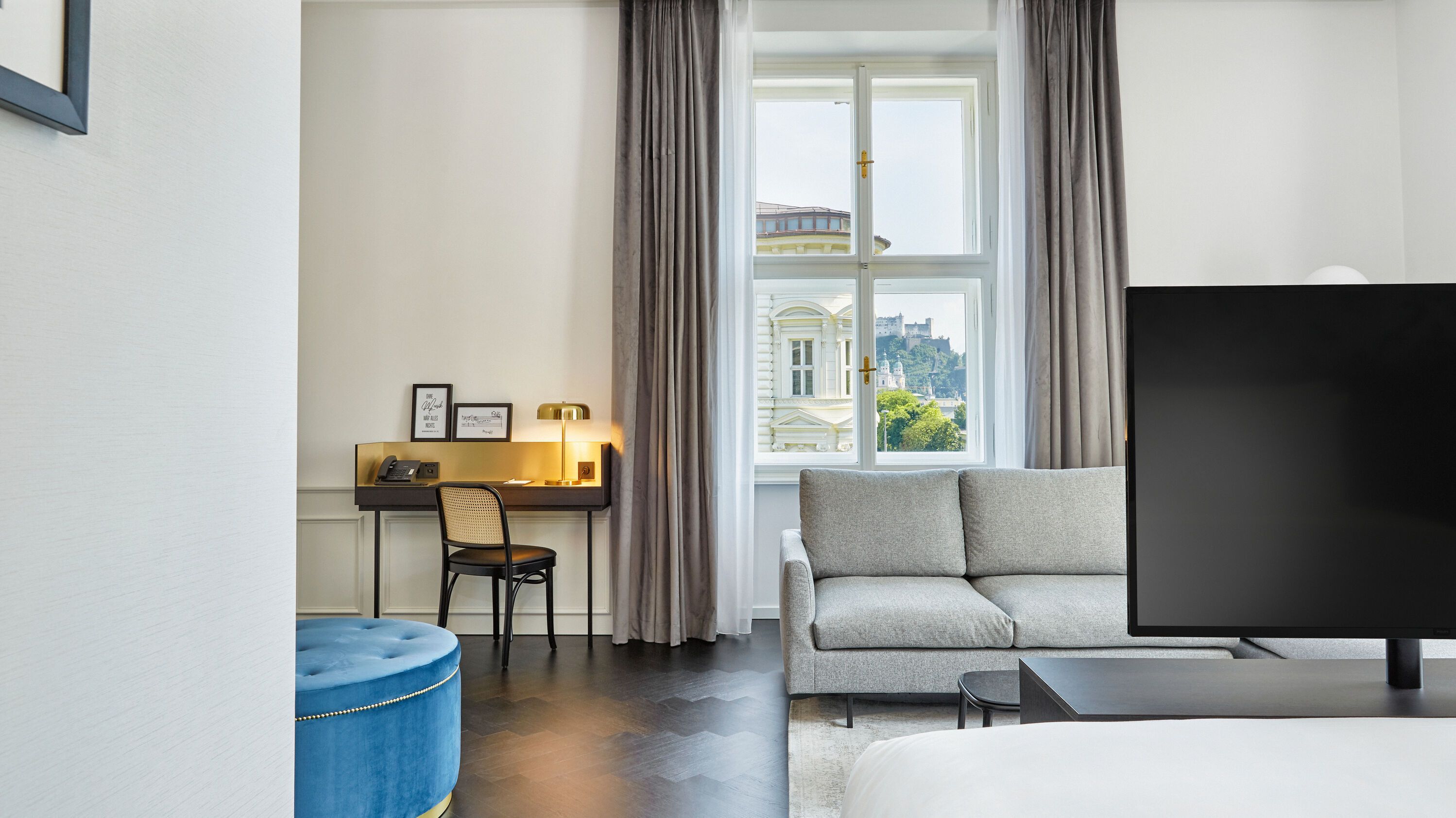 Helles Hotelzimmer mit gemütlichem Sofa, großem Fenster mit Blick auf historische Gebäude, Schreibtisch mit Lampe und Stuhl sowie modernem Flachbildfernseher für entspanntes Arbeiten und Erholen.